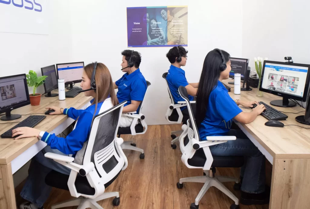 Four InsBOSS virtual assistants wearing blue company shirts and headsets, sitting at their individual, dual-monitor workstations in a clean, modern office.