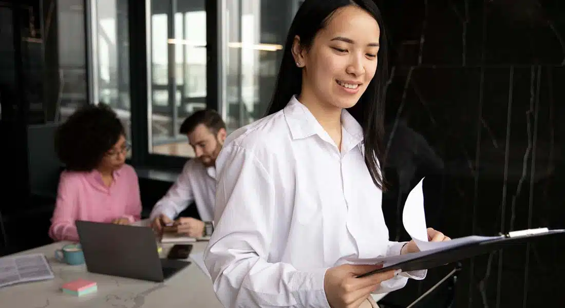 A professional woman smiling while holding a clipboard and looking at documents, with two colleagues working in the background.