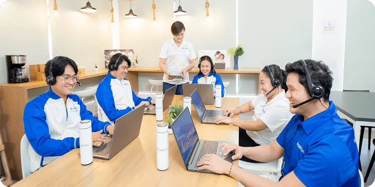 A cheerful team of five virtual assistants wearing matching blue shirts and headsets, working on laptops at a long conference table, supervised by a manager standing behind them.