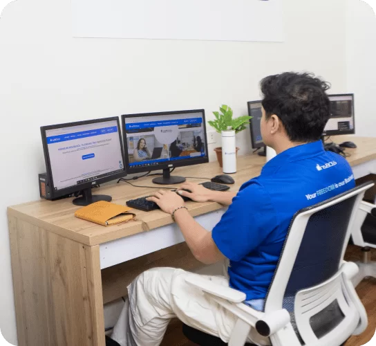 An InsBOSS virtual assistant wearing a blue shirt and headset, working diligently at a desk with a dual-monitor setup.