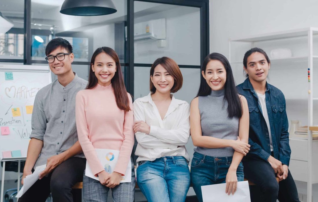 Group of Asia young creative people in smart casual wear looking at camera and smiling in creative office workplace. Diverse Asian male and female stand together at startup. Coworker teamwork concept.