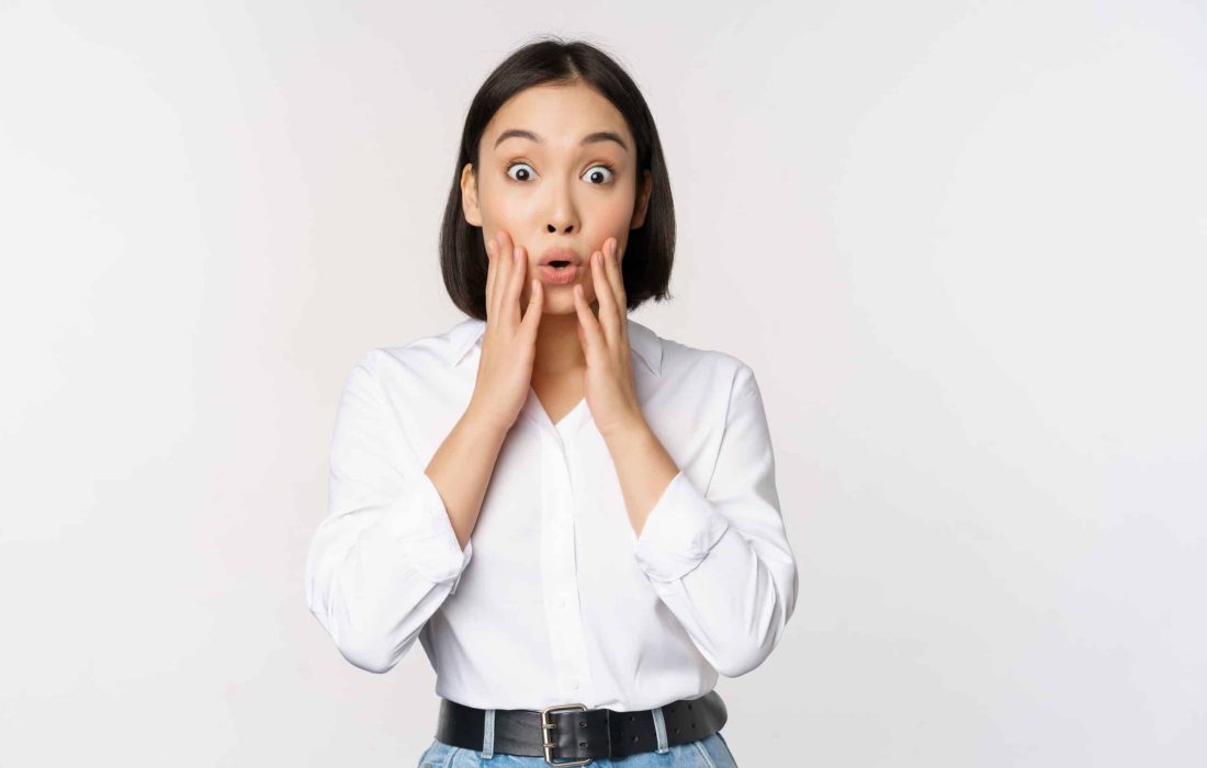 Image of asian female model looking surprised, staring amazed, reacting at surprise big news, standing over white background.