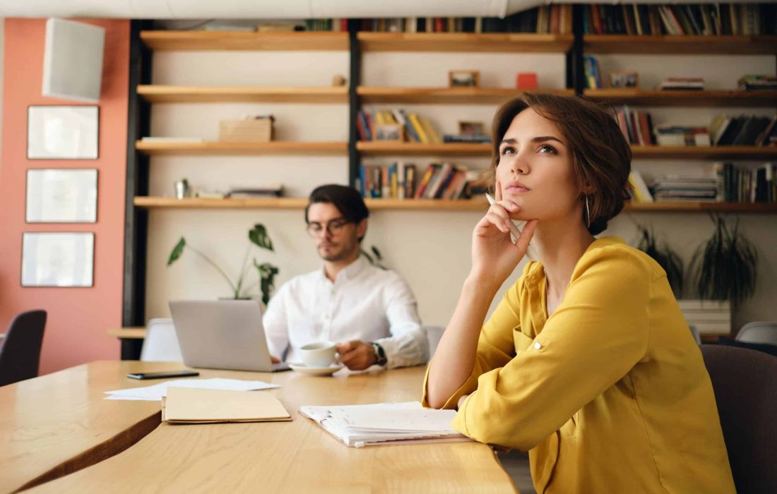 Young pensive woman sitting at the table with notepad dreamily looking aside working with colleague on background in modern office